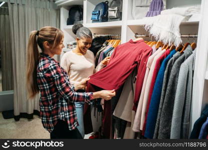 Two girlfriends choosing clothes in shop, shopping. Shopaholics in clothing store, consumerism lifestyle, fashion
