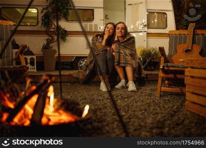 Two girlfriends wrapped in plaid sitting by the campfire in the night, picnic at camping in the forest. Summer adventure on rv, camping-car on background. Girls leisures, travelling with trailer. Girlfriends wrapped in plaid sitting by campfire