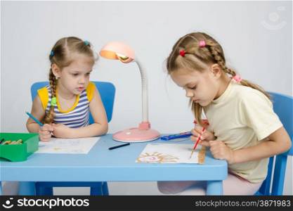 Two girls of four and six years of sitting at the table and draws and paints and pencils. Girl watching sister draws paints
