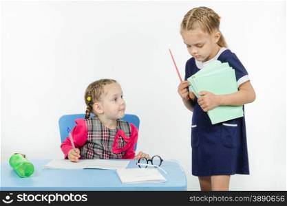 Two girls play school teacher and student. Teacher seeks pupil notebook pile of notebooks