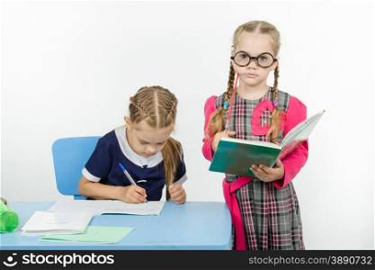 Two girls play school teacher and student. The student writes in a notebook under supervision of teacher
