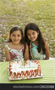 Two girls sitting in front of a birthday cake