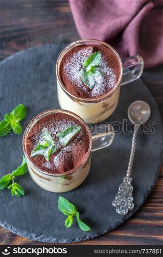 Two glass mugs of tiramisu decorated with cocoa and fresh mint leaves