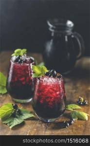Two glass of cold ice black currant juice or cocktail with ripe berries and green leaves on dark wooden table. Alcohol or non alcohol summer fresh drink.