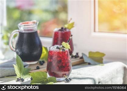 Two glass of cold ice black currant juice with ripe berries and green leaves on table in sunny room near window with garden outside.