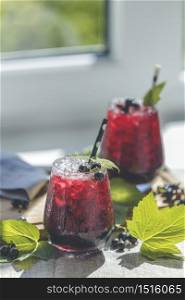 Two glass of cold ice black currant juice with ripe berries and green leaves on table in sunny room near window with garden outside.