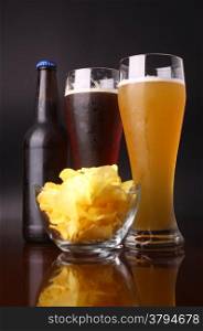 Two glasses of beer, bottle and potato chips over a dark background