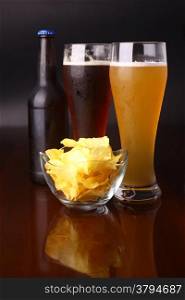 Two glasses of beer, bottle and potato chips over a dark background