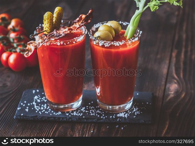Two glasses of Bloody Mary garnished with gherkins, fried bacon strips, green olives and celery stalk
