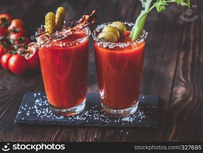 Two glasses of Bloody Mary garnished with gherkins, fried bacon strips, green olives and celery stalk