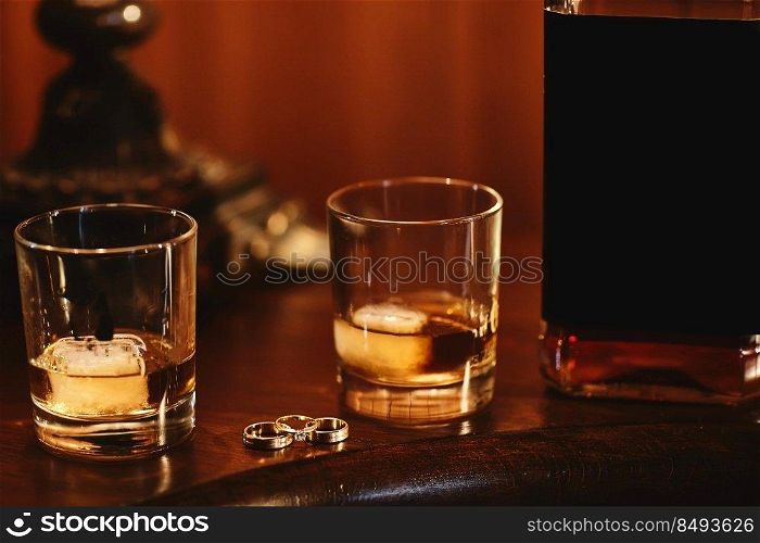 two glasses of cognac with ice, bottle with mockup and gold wedding rings on a wooden table. wedding day.. two glasses of cognac with ice, bottle with mockup and gold wedding rings on a wooden table. wedding day