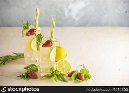 Two glasses of cold icy refreshing drink with lemon and strawberry served with bar tools on light pink table. Fresh cocktail drinks with ice fruit and herb decoration.