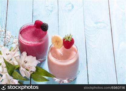 Two glasses of fresh healthy fruit smoothies. Breakfast table with a glass of fresh banana and strawberry smoothie, one with raspberry and wild berries, near white flowers on a blue background.