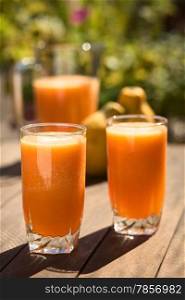 Two glasses of freshly prepared papaya juice with pitcher and papaya fruits in the back on table outdoors (Selective Focus, Focus on the front rim of the first glass)