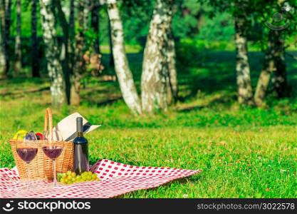 two glasses of red wine and a picnic basket with fruit on a tablecloth in a summer park