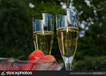 Two glasses with champagne outdoors and a blurred bowl with strawberries in the foreground