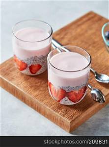 Two glasses with smoothie and pieces of srtawberries, chia seeds on a wooden board on a gray background. Concept of healthy dieting food.. Layered yogurt, homemade dessert with ripe strawberries, chia pudding in glass on a wooden background