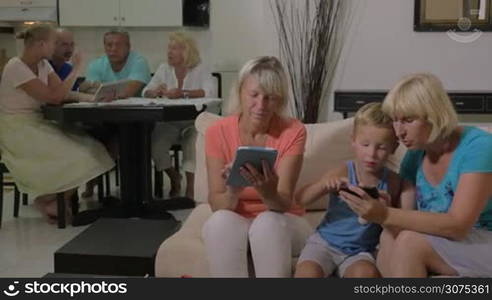 Two grandmothers and little grandchild at home. One woman with a boy playing on smart phone while another grandma and other family members using tablet computers