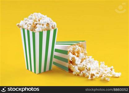 Two green white striped carton buckets with tasty cheese popcorn, isolated on yellow background. Box with scattering of popcorn grains. Movies, cinema and entertainment concept.