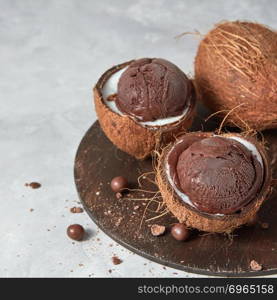 Two halves of coconut with homemade chocolate dessert on a wooden board on a gray background with copy space.. Summer concept.. Tasty fresh brown ice cream in a coconut peel with chocolate balls on a round board on a gray.