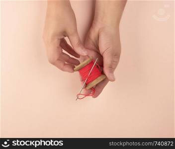 two hands hold a wooden bobbin with red wool threads and an iron needle, peach background