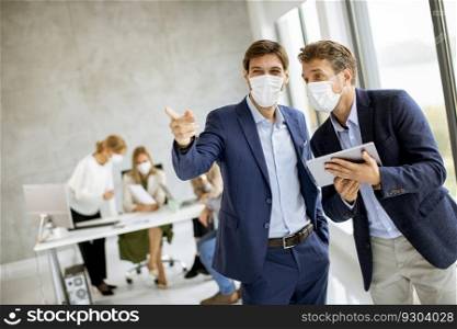 Two handsome young business men with facial protective masks discussing with digital tablet in the office