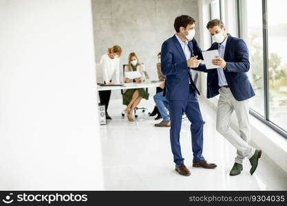 Two handsome young business men with facial protective masks discussing with digital tablet in the office