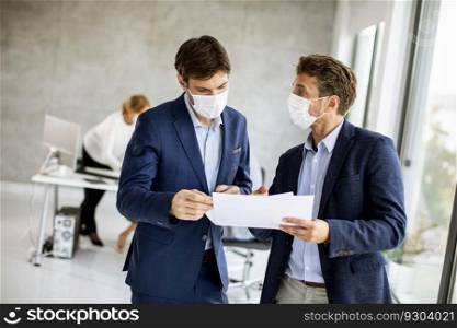 Two handsome young business men with facial protective masks discussing with paper plan in the office