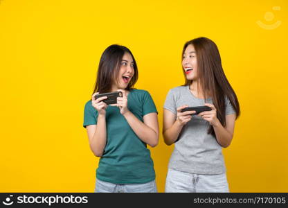 Two Happiness Asian smiling young woman gamer using smart mobile phone and playing games on isolated yellow color background, Lifestyle and leisure with hobby concept