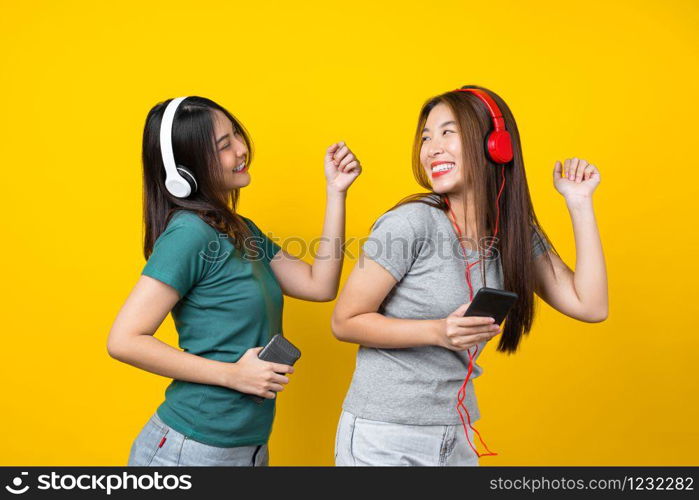 Two Happiness Asian smiling young woman wearing wireless headphones for listening music via smart mobile phone and dancing on isolated yellow color background, Lifestyle and leisure with hobby concept