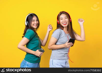 Two Happiness Asian smiling young woman wearing wireless headphones for listening music via smart mobile phone and dancing on isolated yellow color background, Lifestyle and leisure with hobby concept