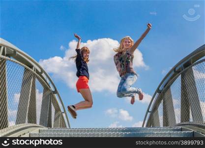 Two happy and enthusiastic teenage girls jumping on bridge in front of blue sky