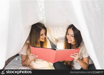 two happy female friends looking album sitting bed white curtain