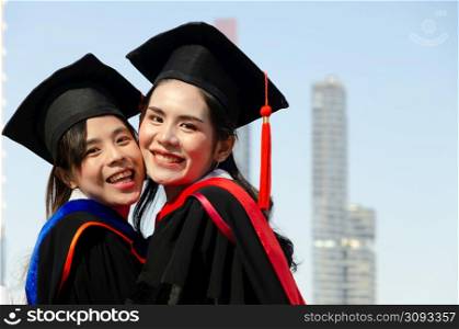 Two happy female in graduation gowns hugging and smiling while standing outdoors with city buildings background