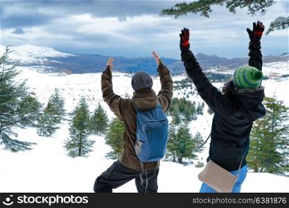 Two happy friends enjoying winter holidays, man and woman standing with raised up hands in the mountains, enjoying landscape covered with snow, having fun on winter vacation