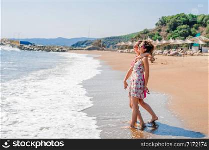 Two happy girls female friends or sisters standing by the seaside on the beach by the sea or the ocean looking to the water smiling wearing summer dress and sunglasses having fun