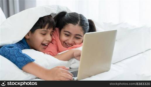 Two happy Indian brother and sister in traditional clothing lying on bed under blanket smiling, using laptop, having fun together at home. Education, Siblings relationship concept