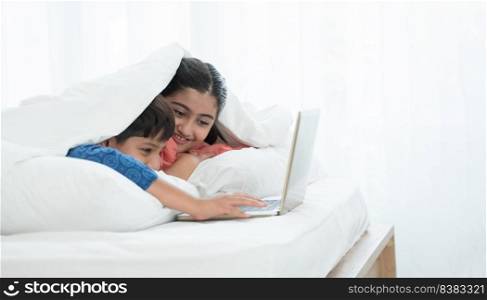 Two happy Indian brother and sister in traditional clothing lying on bed under blanket smiling, using laptop, having fun together at home. Education, Siblings relationship concept. Selective focus