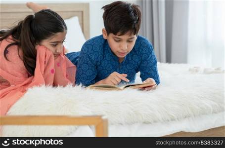Two happy Indian brother and sister in traditional clothing lying on bed and reading book, having fun together at home. Education, Siblings relationship concept