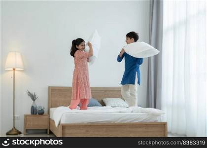 Two happy Indian brother and sister in traditional clothing standing on bed, playing pillow fight, having fun together at home. Playful kids, Siblings relationship concept