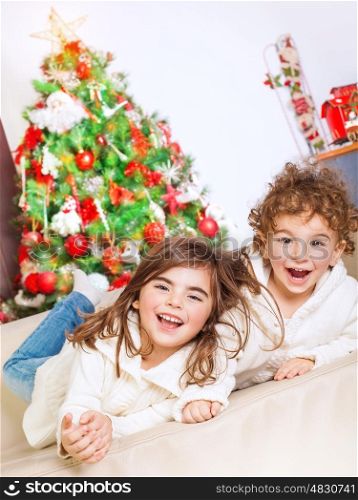 Two happy kids lying down on sofa near beautiful decorated Christmas tree and laughing, cheerful family spending winter holidays at home