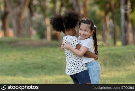 Two happy little diverse kids, African and Caucasian girls hugging each other while playing together in green park. Diversity ethnic friendship concept