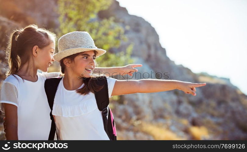 Two happy little girls enjoying excursion to the mountains, with pleasure discovering wild nature, with wonder looking on something and pointing, happy active time in a summer camp