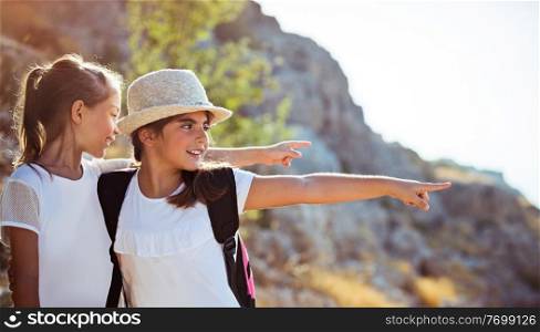 Two happy little girls enjoying excursion to the mountains, with pleasure discovering wild nature, with wonder looking on something and pointing, happy active time in a summer camp