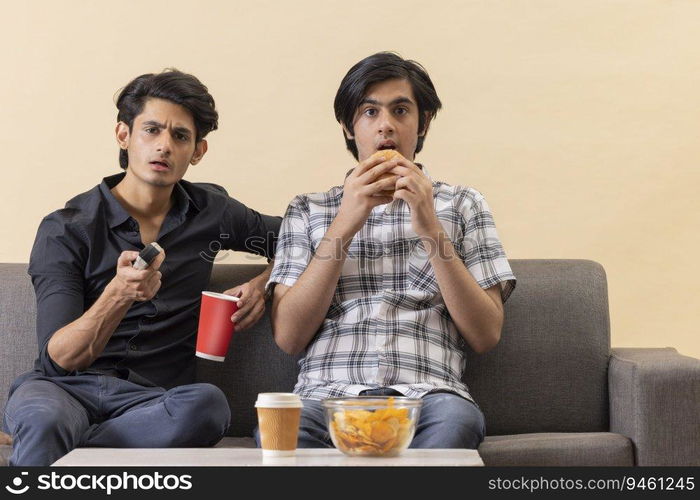 Two happy teenage boys eating fast foods and watching TV together at home