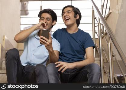 Two happy teenage boys using smartphone while sitting on stairs at home