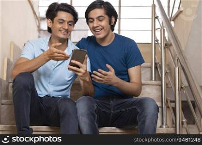 Two happy teenage boys using smartphone while sitting on stairs at home