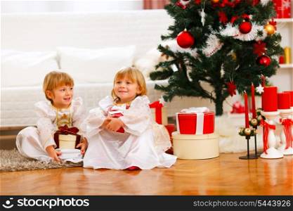 Two happy twins girl sitting with gift boxes near Christmas tree