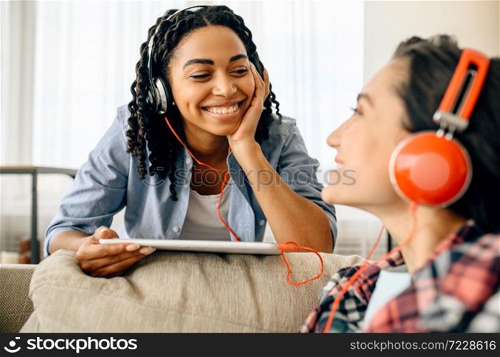 Two happy women enjoys listening to music at home. Pretty girlfriends in earphones relax in the room, sound lovers resting on couch, female friends leisures together. Two happy women enjoys listening to music at home