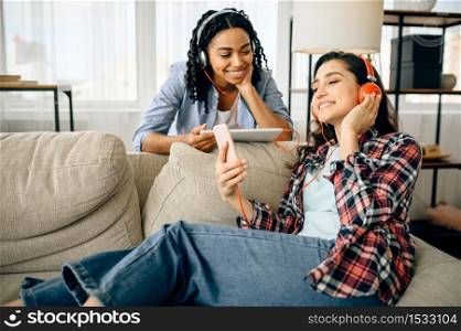 Two happy women enjoys listening to music indoors. Pretty girlfriends in earphones relax in the room, sound lovers resting on couch, female friends leisures together. Happy women enjoys listening to music indoors